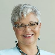 A smiling female-presenting person with short, gray hair wearing a pearl necklace against a plain brown background