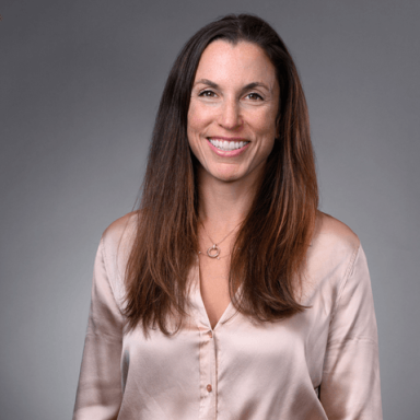 A smiling female-presenting person with long brown hair wearing a silky peach top against a gray background.