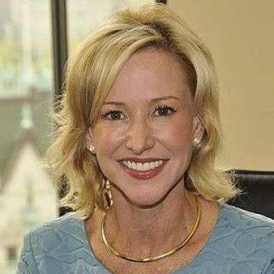 A smiling female-presenting person with blonde hair wearing a gold necklace against a blurred background.