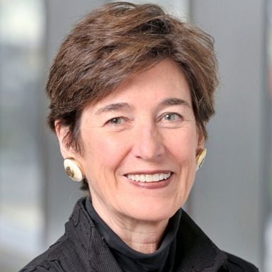 A smiling female-presenting person with short brown hair wearing a black top against a blurred background.