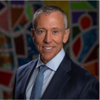 Smiling light-skinned, male-presenting individual with short greying hair wearing a suit with artwork backdrop