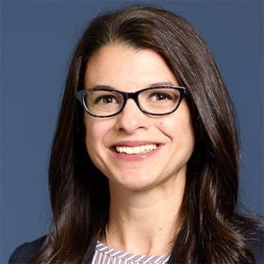 Smiling light-skinned, female-presenting individual with long dark hair wearing a dark shirt on a bkue backdrop.       