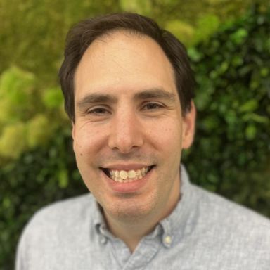 Smiling light-skinned, male-presenting individual with short dark hair wearing a grey shirt on an outdoor backdrop.