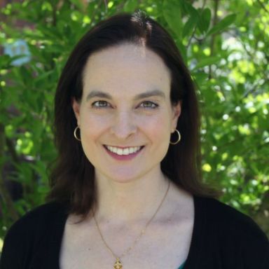 Smiling light-skinned, female-presenting individual with long dark hair wearing a dark shirt on an outdoor backdrop.