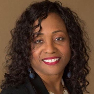 A smiling dark-skinned female with black hair wearing a dark suit jacket against a brown background