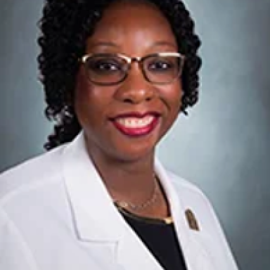 A smiling darker-skinned female with black curly hair wearing glasses and a lab coat against a light gray backgroundt