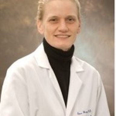 A smiling light-skinned female with blonde hair wearing a black turtleneck under a lab coat against a brown marbled background