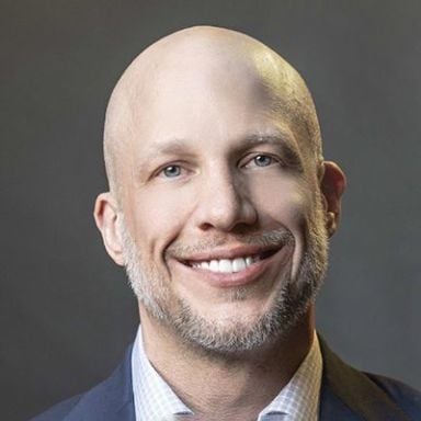 A smiling light-skinned male with no hair and a short gray beard wearing a navy suit jacket against a dark gray background