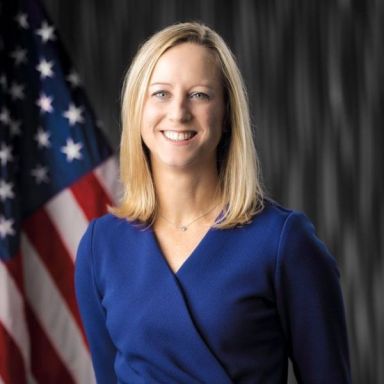 A smiling light-skinned female with blonde hair wearing a navy shirt against a gray background with an American flag