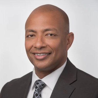 A smiling darker-skinned male wearing a dark gray suit and tie against a white background