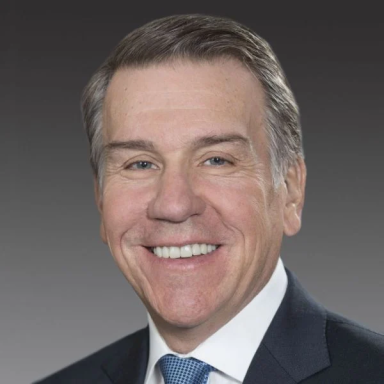 A smiling light-skinned male with gray hair wearing a dark suit and blue tie against a gray gradient background