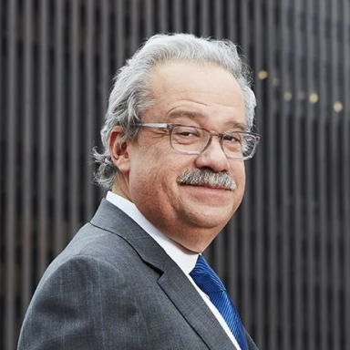 A smiling light-skinned male with gray hair and a mustaches wearing glasses and a gray suit and tie against an ambiguous background