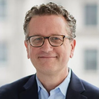 A smiling light-skinned male with gray hair wearing glasses and a navy suit jacket over a blue button-up shirt against an ambiguous background