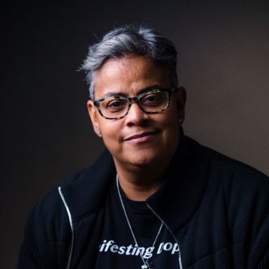 A smiling darker-skinned individual with gray hair wearing glasses and a black graphic t-shirt and a black jacket against a dark gray background