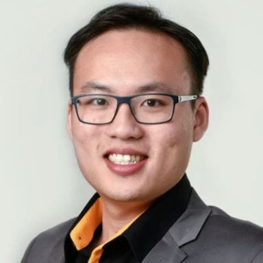 A smiling male of Asian heritage with black hair wearing glasses and a gray suit jacket over a black shirt against a white background