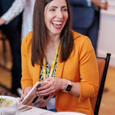 A smiling light-skinned female with brown hair wearing a yellow blazer over a patterned shirt against an ambiguous background