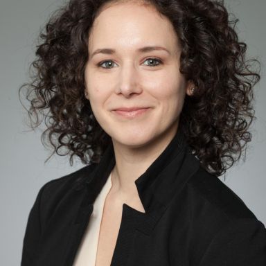 A woman with short, curly hair in a black blazer against a gray background.