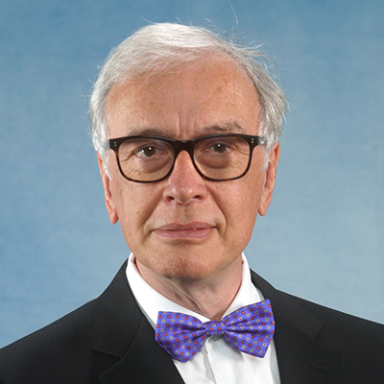 A light-skinned male with short white hair wearing a black suit and bowtie and glasses against a blue background