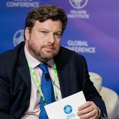 A male conference presenter against the the Milken Institute Global Conference backdrop.