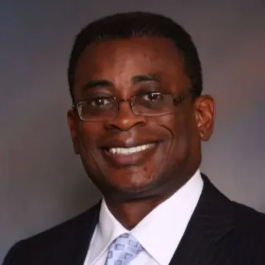 A smiling Black man wearing a dark suit and tie and glasses against a gray background