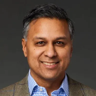 A smiling male of Indian heritage with black and gray hair wearing a tan jacket of a button-up shirt against a dark gray background