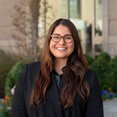 An image of an adult woman named Nadia Sarfraz. She has tan white skin, long brown hair, and she is wearing glasses and a black jacket and smiling at the camera.