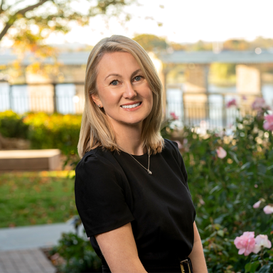 Smiling light-skinned female-presenting person with light hair wearing a black shirt against a nature-ish background