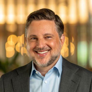 A headshot of a man named Peter Mintzberg who is a Fintech Advisory member of the Milken Institute. He is smiling and has a slight beard and short brown/gray hair, white skin, and is wearing a suit.
