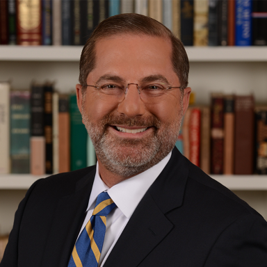 An image of a man named Alex Azar. He is standing in front of a bookshelf and smiling at the camera. He is middle-aged in appearance, has white skin, short brown hair, and a gray-ish brown stubbled beard. He is wearing a suit jacket over a white collared shirt with a blue and yellow striped tie, and round glasses.
