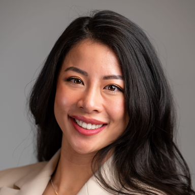 A headshot of a woman named Angelina Fung. She is smiling at the camera and standing against a gray background. She has long black hair, brown eyes, and is Asian in appearance with white skin and wearing a beige blazer.