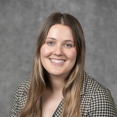 A headshot of a woman named Ainslie Tisdale. She has white skin, long brunette-ish blonde-ish hair, and blue eyes. She is smiling at the camera and wearing a checkered blazer, standing against a gray background.