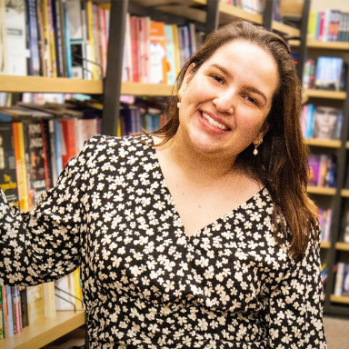 A woman named Michelle Rincon smiles warmly while standing in front of bookshelves in a bookstore or library. She is wearing a black dress with a white floral pattern and gold earrings, with her brown hair down and slightly parted to the side.