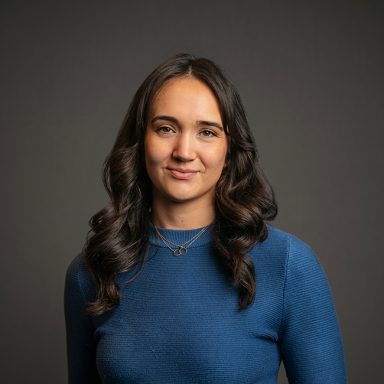 Professional headshot of Abigail Humphreys, a woman with long dark brown wavy hair, wearing a blue ribbed top and a gold necklace. She is smiling softly and posed against a dark gray background.