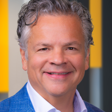 Alberto Casellas, a middle-aged man with short, wavy gray hair, wearing a blue textured blazer over a white dress shirt, smiling in front of a blurred background with yellow, gray, and black vertical elements.