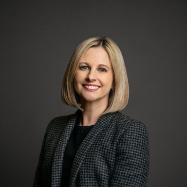 Professional headshot of Belle Tabas, a woman with straight blonde hair in a sleek bob, wearing a black and white houndstooth blazer over a black top. She is smiling and posed against a dark gray background.
