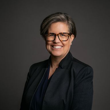 Professional headshot of Brenda Huneycutt, a woman with short gray hair and tortoiseshell glasses, wearing a dark textured blazer over a navy blouse. She is smiling broadly against a dark gray background.