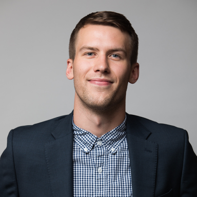 Professional headshot of Cameron McConkey, a man with short light brown hair, wearing a navy blazer over a blue and white checkered button-down shirt. He is smiling slightly and posed against a light gray background.