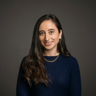 Professional headshot of Christina Dialynas, a woman with long dark brown wavy hair, wearing a navy ribbed top and a gold chain necklace. She is smiling gently and posed against a dark gray background.