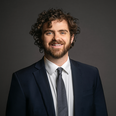 Professional headshot of Christopher Multerer, a man with curly dark brown hair and a beard, wearing a dark navy suit, white shirt, and gray tie. He is smiling and posed against a dark gray background.