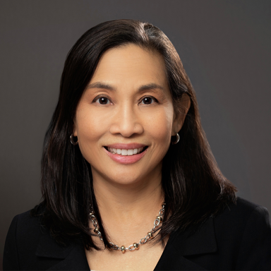 Professional headshot of Diane Ty, a woman with straight black hair and a warm smile, wearing a black blazer and a silver chain necklace. She is posed against a neutral dark background.