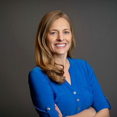 Professional headshot of Erika Stratmann, a woman with long blonde hair styled in loose waves, wearing a vibrant blue blouse with rolled sleeves. She is smiling with arms crossed and posed against a dark gray background.