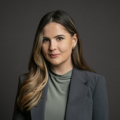 Professional headshot of Gabby Hines, a woman with long brown hair styled in loose waves, wearing a soft green top and a charcoal blazer. She is posed against a dark gray background with a slight smile.