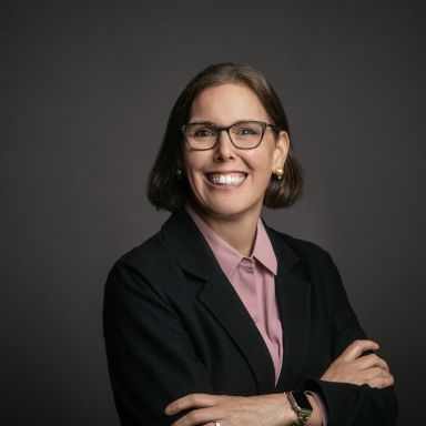 Professional headshot of Holly Freishtat, a woman with short brown hair wearing glasses, pearl earrings, a mauve collared shirt, and a black blazer. She is smiling with arms crossed and posed against a dark gray background.