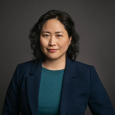 Professional headshot of June Cha, a woman with short curly black hair, wearing a teal top and navy blazer. She is posed against a dark gray background with a confident expression.