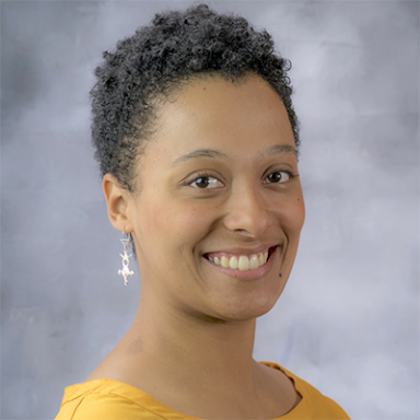 Headshot of a smiling woman with short curly hair, wearing a mustard yellow top and dangling earrings, set against a soft gray studio backdrop.