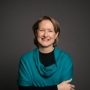 Professional headshot of Kristin Schneeman, a woman with short light brown hair, wearing a teal cowl-neck sweater over a black top. She is smiling warmly with arms loosely crossed, posed against a dark gray background.