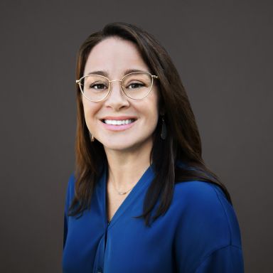 Professional headshot of Lauren Dunning, a woman with straight brown hair wearing round gold glasses, drop earrings, and a cobalt blue blouse. She is smiling and posed against a dark gray background.