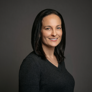Professional headshot of Lena Flaum, a woman with straight dark brown hair, wearing a charcoal V-neck top and a small pendant necklace. She is smiling slightly and posed against a dark gray background.