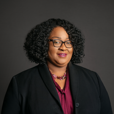 Professional headshot of Lisa Lewis, a woman with short curly black hair and glasses, wearing a dark blazer over a burgundy blouse and a pink beaded necklace. She is smiling softly and posed against a dark gray background.
