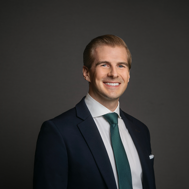 Professional headshot of Mike Brown, a man with slicked-back blonde hair, wearing a dark navy suit, white shirt, and teal tie. He is smiling and posed against a dark gray background.